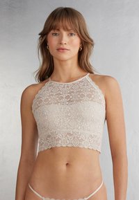 Bralette de encaje en beige claro con bordes festoneados, que presenta intrincados patrones florales y un diseño de cuello halter. Textura suave, forma ajustada.