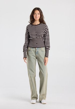 Femme debout sur un fond uni portant un pull rayé noir et blanc, un jean large vert clair et des baskets blanches.