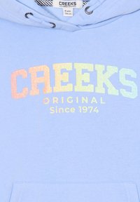 Sudadera azul claro con un bolsillo frontal que presenta un estampado brillante multicolor que dice "CREEKS ORIGINAL Desde 1974". Hecha de una tela suave.