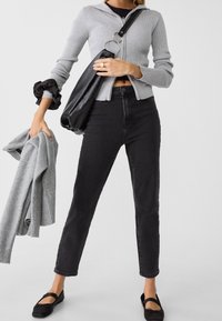 Cárdigan gris con cremallera y costuras, jeans negros de talle alto, bolso negro de hombro, sosteniendo suéter gris. Zapatos planos negros completan el conjunto.