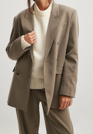 Femme portant un blazer oversize taupe et un pantalon assorti sur un pull en maille blanc, la main tenant le revers du blazer.