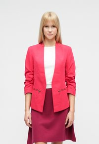 Blazer aderente rosa acceso con collo a scialle e due tasche frontali con cerniera, abbinato a un top bianco e una gonna magenta.