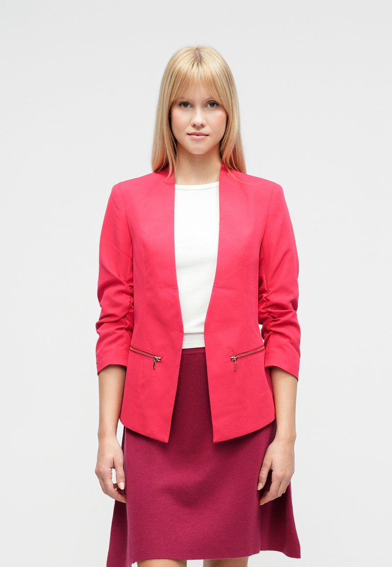 Blazer aderente rosa acceso con collo a scialle e due tasche frontali con cerniera, abbinato a un top bianco e una gonna magenta.