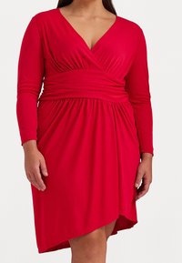 Červené šaty s dlouhými rukávy, výstřihem do V a nařaseným pasem. Měkká látka splývá v A-linii s vysokým a nízkým lemem.