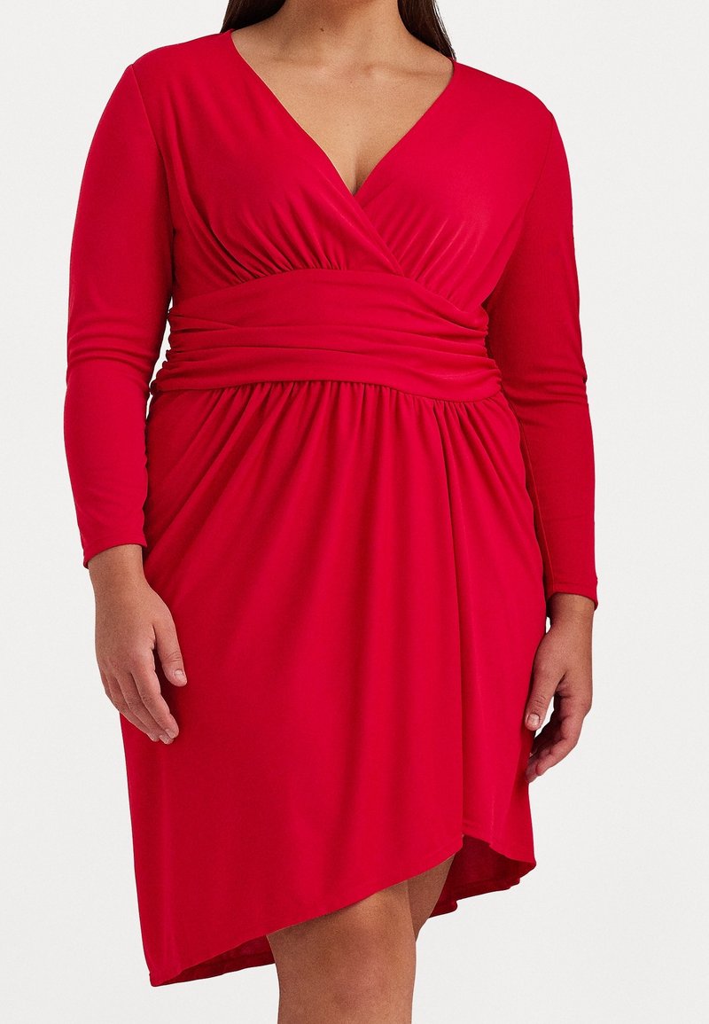 Červené šaty s dlouhými rukávy, výstřihem do V a nařaseným pasem. Měkká látka splývá v A-linii s vysokým a nízkým lemem.