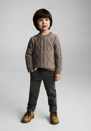 Brun kabelstickad tröja med rund halsringning, grå jeans och beige snörstövlar. Barnet står självsäkert med händerna i fickorna.