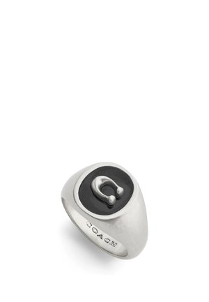 Anillo de plata con un centro ovalado negro y un icónico símbolo metálico prominente. Diseño suave y redondeado con el detalle grabado "COACH".
