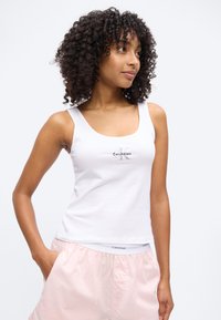 Νέα γυναίκα με σγουρά μαλλιά που φοράει λευκό φανελάκι Calvin Klein και ανοιχτό ροζ σορτς, στέκεται με το ένα χέρι στην τσέπη.