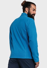 Niebieska polarowa kurtka z wysokim kołnierzem, fakturowanym materiałem i płaskimi szwami, w zestawie z ciemnymi spodniami. Widok z tyłu ukazuje dopasowany krój.