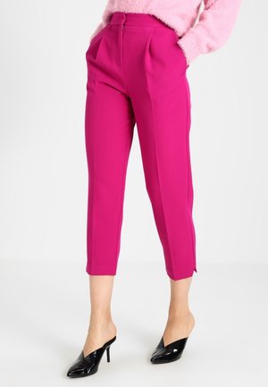 Mujer vestida con pantalones de sastre cortos de color rosa brillante, un suéter rosa peludo y mules negros de tacón alto y punta, posando frente a un fondo blanco.