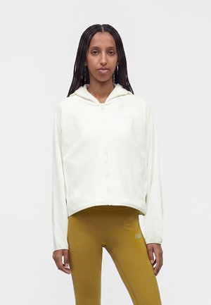 Donna con capelli lunghi intrecciati che indossa una giacca bianca con cappuccio e zip e leggings giallo senape, in piedi davanti a uno sfondo semplice.