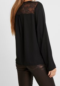 Czarna bluzka z długim rękawem z koronkowym przodem, zwiewnym tyłem i gładkiego materiału. Połączona z błyszczącymi, ciemnobrązowymi spodniami.