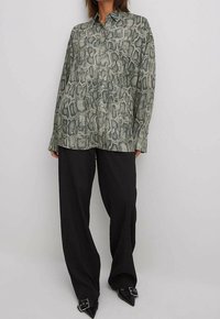 Langermige shirt met een groene en grijze slangenprint, kraagontwerp en knoopsluiting, gecombineerd met brede zwarte broeken en puntige schoenen.