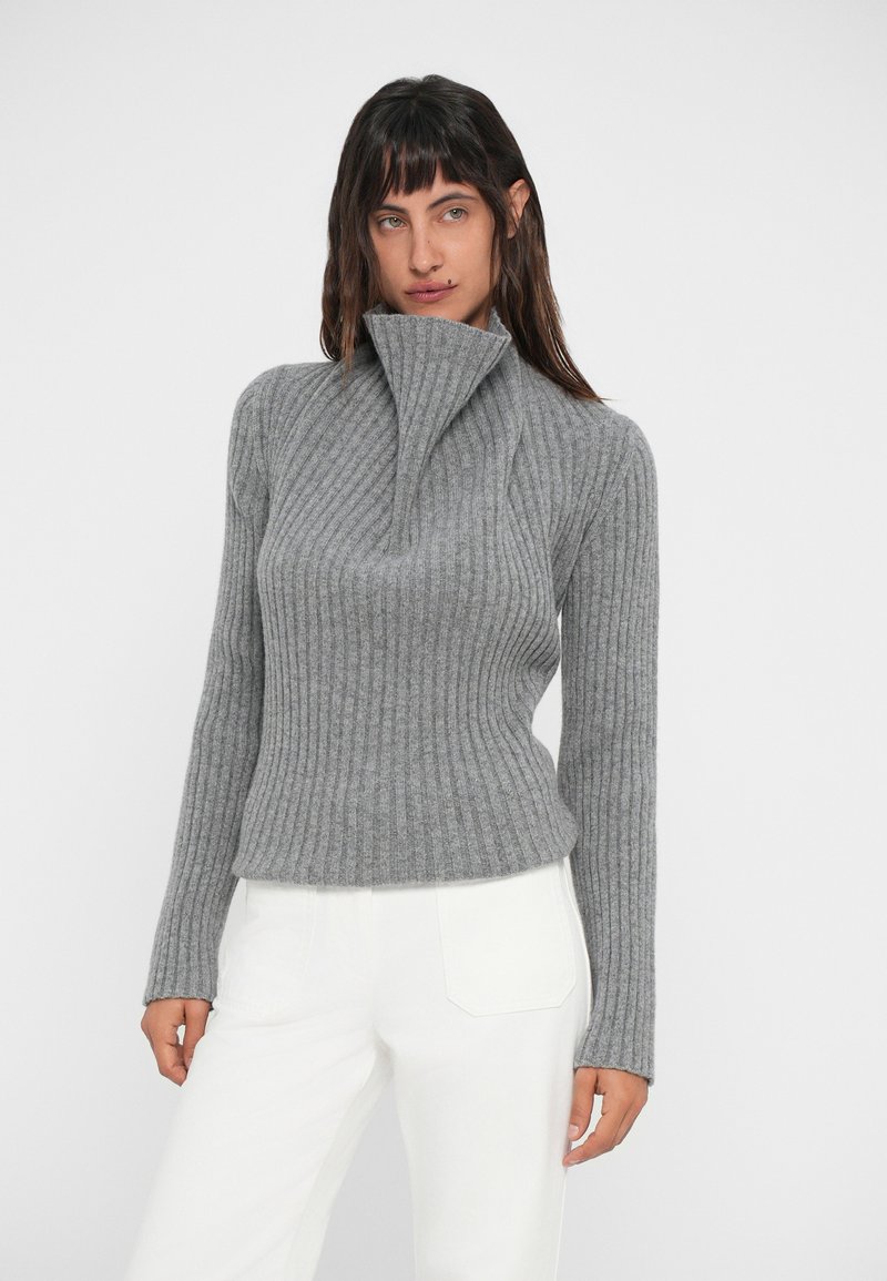 Grey ribbed turtleneck sweater with a high collar, long sleeves, and a fitted silhouette, paired with white pants. Soft texture with vertical stripes.