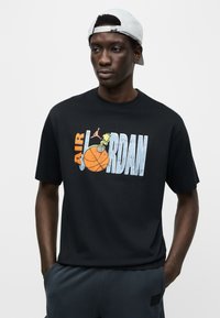 Μαύρο βαμβακερό μπλουζάκι με έντονο, πολύχρωμο γραφικό "AIR JORDAN" που περιλαμβάνει μια μπάλα μπάσκετ και εκρηκτικά σχεδιαστικά στοιχεία. Κοντά μανίκια και στρογγυλή λαιμόκοψη.