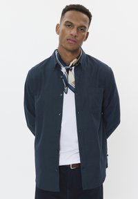 Camisa de botón navy de manga larga, con bolsillo en el pecho, combinada con una bufanda de patrón crema y azul, camiseta blanca debajo, llevada con pantalones oscuros.