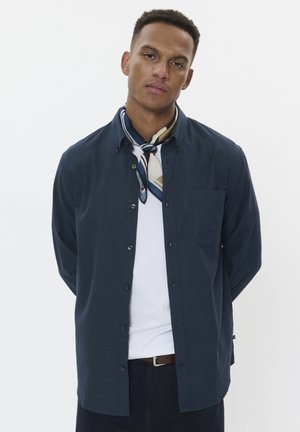 Camisa de botón navy de manga larga, con bolsillo en el pecho, combinada con una bufanda de patrón crema y azul, camiseta blanca debajo, llevada con pantalones oscuros.