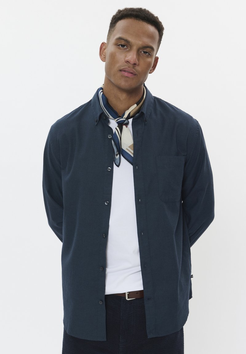 Camisa de botón navy de manga larga, con bolsillo en el pecho, combinada con una bufanda de patrón crema y azul, camiseta blanca debajo, llevada con pantalones oscuros.