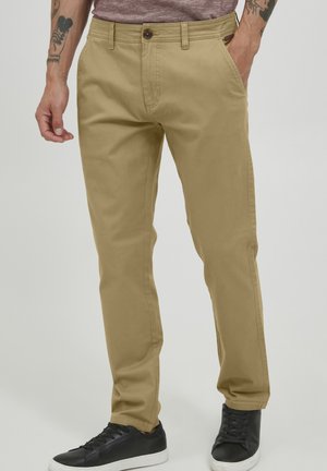 Chinos in cotone beige con una silhouette a gamba dritta, dotati di tasche frontali e chiusura con bottone, indossati con sneakers nere.
