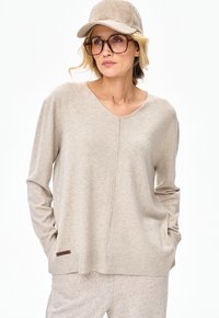 Kobieta w beżowym swetrze z dekoltem w serek, pasujących teksturowanych spodniach, dużych okularach w oprawkach w kolorze żółwia oraz beżowej czapce ze sztruksu, stojąca z rękami w kieszeniach.