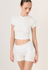 Bílý žebrovaný crop top s kulatým výstřihem a krátkými rukávy, zkombinovaný s bílými přiléhavými šortkami. Na obou kusech se nachází logo "LAMANIA".