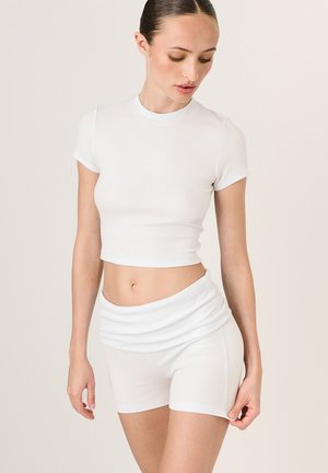 Biely rebrovaný crop top s okrúhlym výstrihom a krátkymi rukávmi, v kombinácii s bielymi priliehajúcimi šortkami. Obe kusy majú logo "LAMANIA".