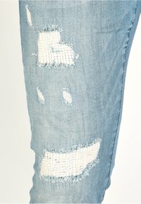 Ljusblå denimjeans med slitna lappar, med fransade kanter och vävt mönster under revorna. Närbild på knäområdet.