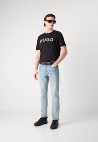 Melns T-krekls ar "HUGO" logotipu, gaiši zili džinsi ar krāsas traipiem, melni sporta apavi un melnas saulesbrilles; modelis neitrāla fona apstākļos.