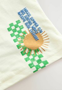 Camiseta de algodón de color amarillo claro con un diseño gráfico de un sol en dorado y marrón, con texto en azul y verde y un patrón a cuadros verdes.