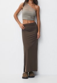 Vrouw in een beige een-schouder crop top, donkerbruine hoog-taille lange rok met een zijnaad, en bruine gespikkelde loafers.