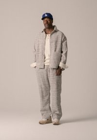 Ensemble assorti en tissu tissé gris avec veste et pantalon, au tissu texturé. Haut en tricot beige clair en dessous. Casquette bleue et baskets beiges complètent le look.