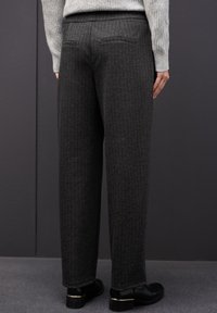 Pantalones grises texturizados de corte recto, con bolsillos traseros. Llevados con zapatos negros, exhibidos contra un fondo oscuro.