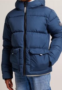 Blå puffjacka med huva, med dragkedjeslutning, två framfickor och rutigt mönster. Tillverkad av ett slätt, isolerat material.