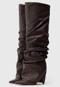 Par de botas altas, holgadas, de color marrón oscuro y textura, con punteras puntiagudas y tacones de cuña, sobre un fondo liso.