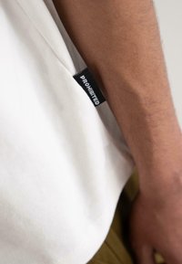 White cotton T-shirt with a smooth texture and a black label reading "PROHIBITED" on the left side, partially obscured by the arm.