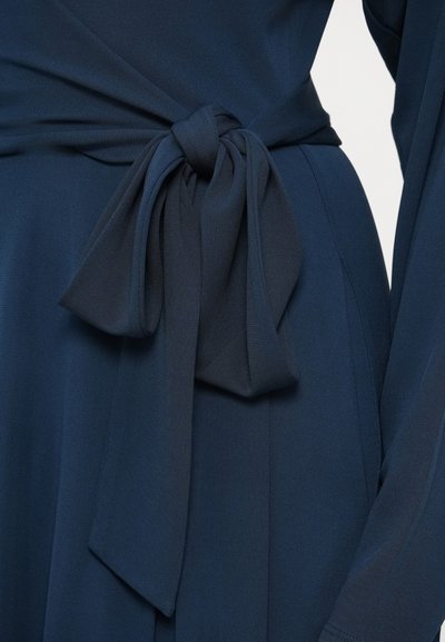 Tumši zila kleita ar mīkstu tekstūru, viduklī ar piesaistāmu jostu un garām, plūstošām piedurknēm.