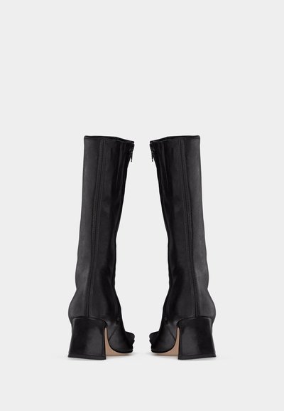 Μαύρες μπότες με τακούνι έως τη μέση της γάμπας, με φερμουάρ στο πλάι, στρογγυλή μύτη και χοντρό τακούνι, εμφανιζόμενες από πίσω σε λευκό φόντο.