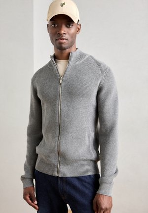 Grå zip-up genser med ribbet tekstur, høy krage og tettsittende ermer, båret over en lys skjorte, parret med mørke denimjeans.