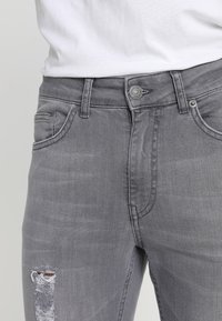 Jean gris de mezclilla con cierre de botón, bolsillos frontales y detalle de rodilla desgastada. Textura suave con un diseño ajustado.