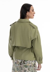Giacca in finta pelle verde oliva, design corto, ampio colletto, spalline sulle spalle e polsini con bottoni, indossata sopra una gonna a fantasia.