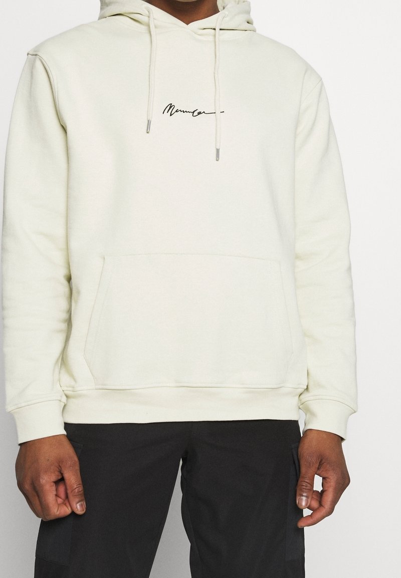 Sudadera crema con cordón, bolsillo frontal y logotipo bordado en negro. Tejido suave con un ajuste relajado. Combinada con pantalones oscuros.