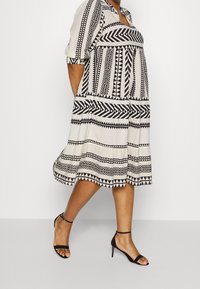 Vestido de patrones en blanco y negro con mangas cortas, cuello redondo y falda acampanada. Combinado con zapatos de tacón alto negros con tiras.
