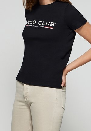 T-shirt en coton noir à manches courtes et col rond. Présente un texte graphique "POLO CLUB" en blanc et rouge avec l'année de création.
