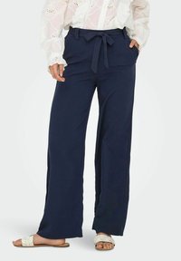 Pantalon large bleu marine à taille nouée, confectionné dans un tissu lisse, porté avec un chemisier blanc à manches longues brodé et des sandales légères.