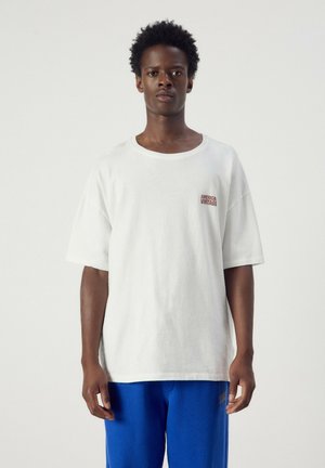 Giovane uomo in piedi, indossa una maglietta bianca oversize con il testo "America Vintage" e pantaloni blu brillanti su uno sfondo chiaro e uniforme.