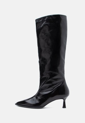 Botte en cuir verni noir, hauteur genou, avec un bout pointu et un petit talon, vue de côté droit sur fond blanc.