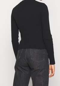 Personne portant un haut noir à manches longues côtelé et un jean en denim gris foncé taille haute, vue de dos.