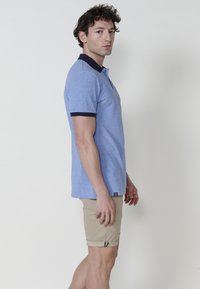 Gaiši zila polo krekls ar teksturētu rakstu, tumši zila apkakle un manšetes, valkāts ar bēšiem šortiem ar sarullētajiem malām un minimālu aparatūru.