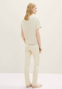 Haut beige clair à manches courtes associé à un pantalon crème à motifs présentant des formes géométriques ; le mannequin se tient pieds nus, tourné vers l'arrière.