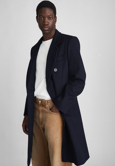 Navy wollen lange jas met een enkele knoop, notch revers en twee zijzakken, gecombineerd met een wit shirt en tan hoge taille jeans.